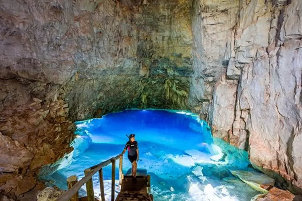 Uma pessoa com roupa de mergulho (snorkeling) está em uma passarela de madeira dentro de uma caverna, olhando para o incrível lago subterrâneo de água azul-turquesa brilhante, ilustrando a principal atração de um roteiro de 7 dias em Bonito, Mato Grosso do Sul.