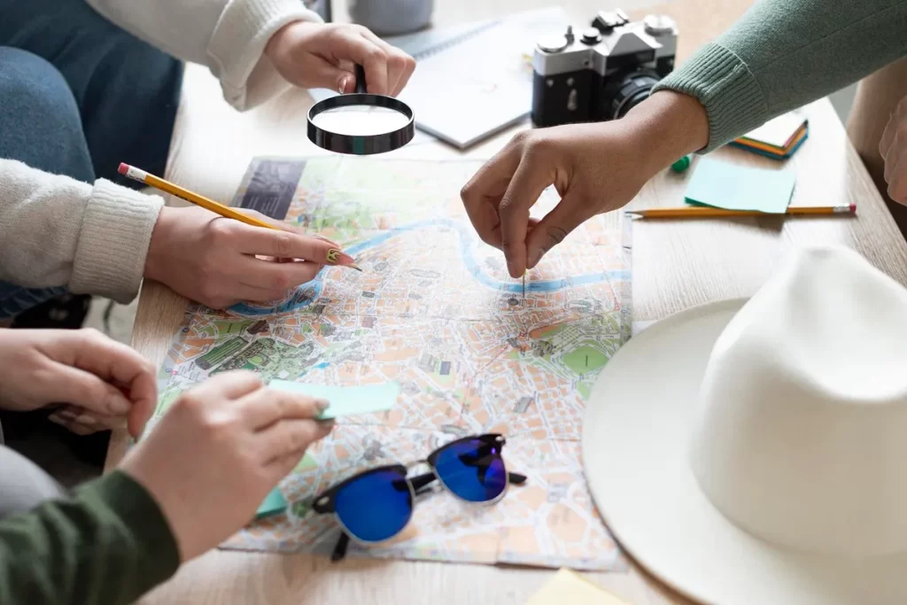 Mãos de quatro pessoas apontando e marcando pontos num mapa de papel sobre uma mesa, ao lado de câmera fotográfica, chapéu e óculos de sol