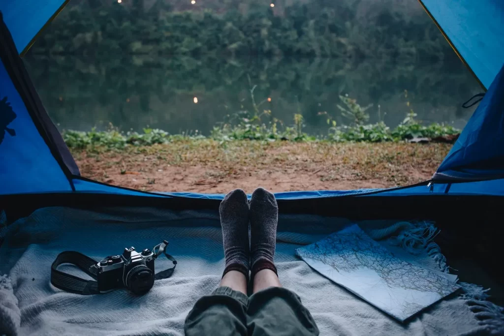 Pessoa relaxando dentro de uma barraca azul, com vista para um lago tranquilo, ao lado de um mapa e uma câmera fotográfica.