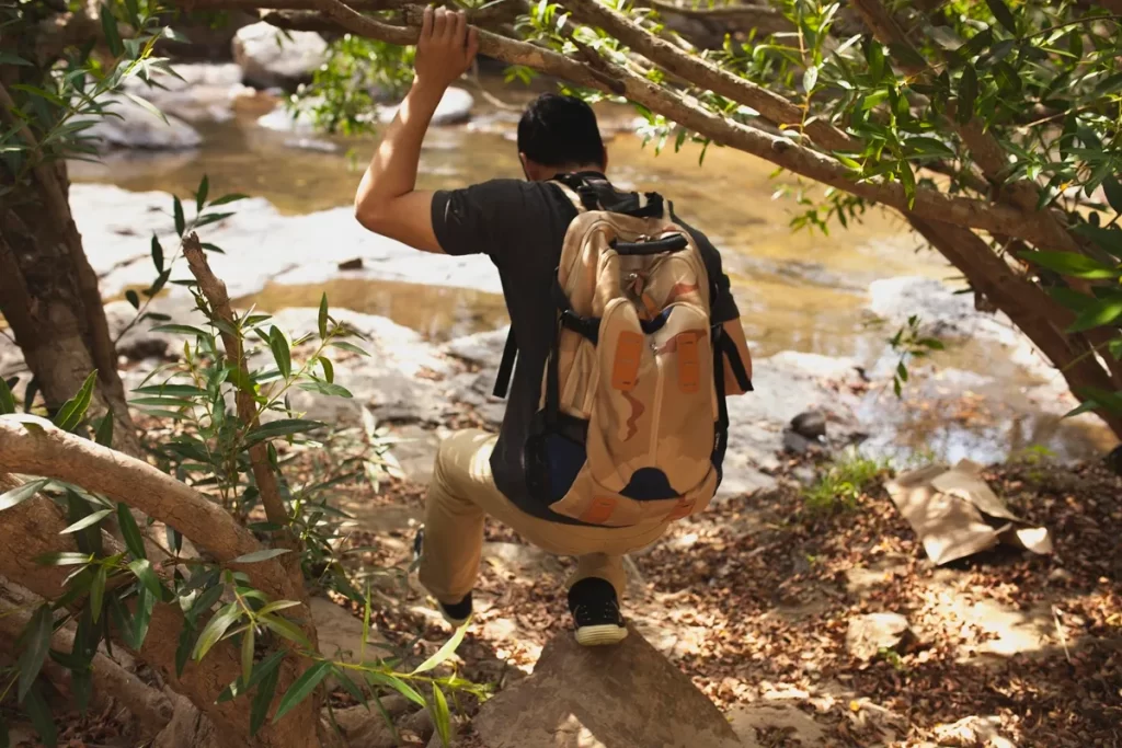 Homem com mochila explora uma trilha ecológica descendo por um caminho em meio à vegetação densa próximo a um rio.