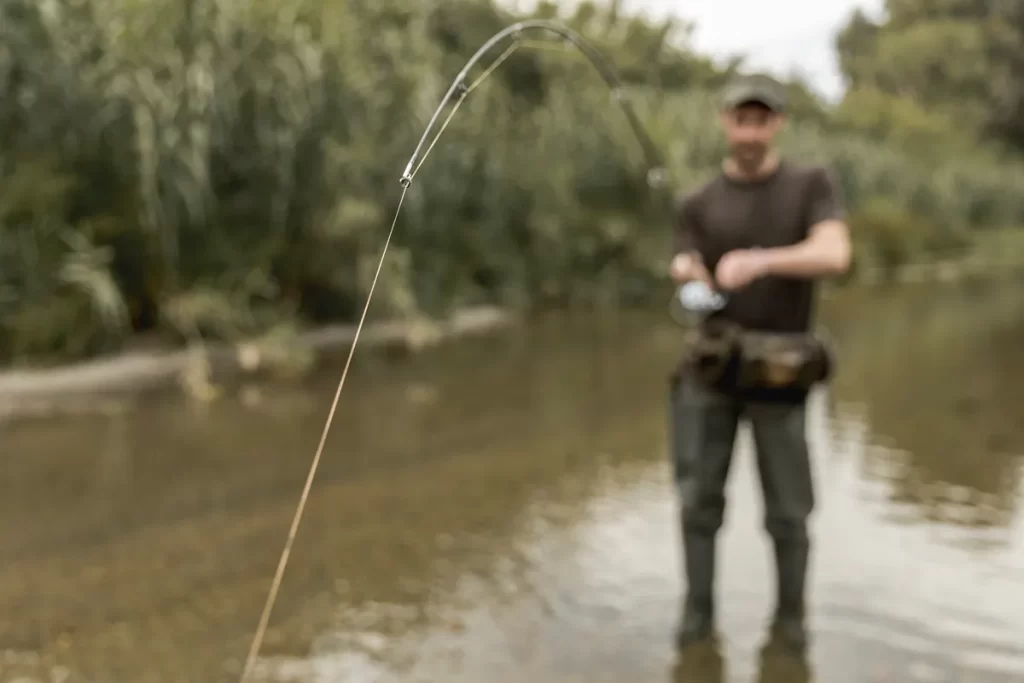 Homem pescando em um rio raso com vara de pesca flexionada, mostrando um momento de captura de peixe.