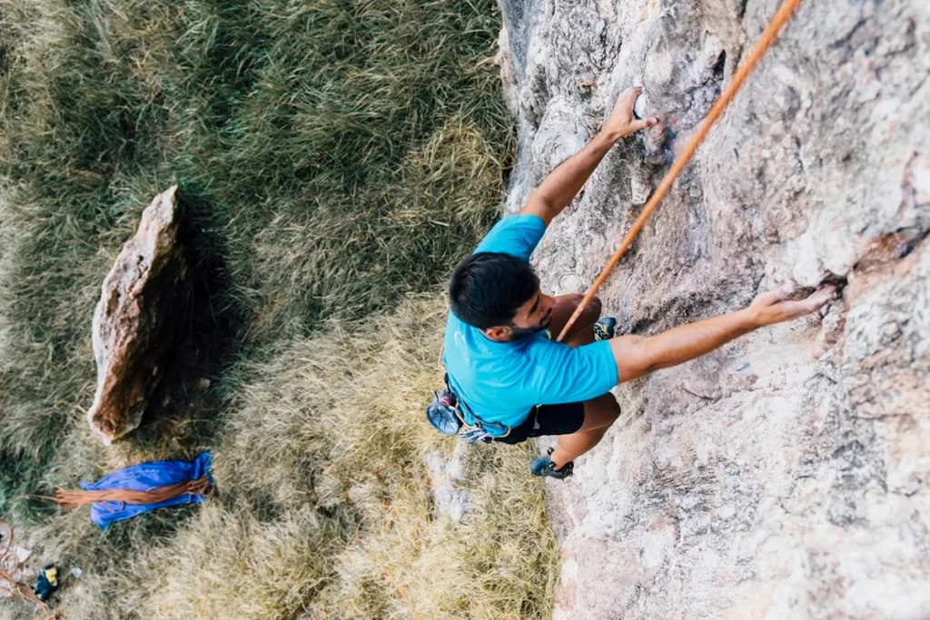 Homem vestindo camiseta azul realiza escalada em uma parede de rocha, utilizando corda de segurança e equipamentos de proteção, enquanto segura firmemente em pequenas fendas da superfície.