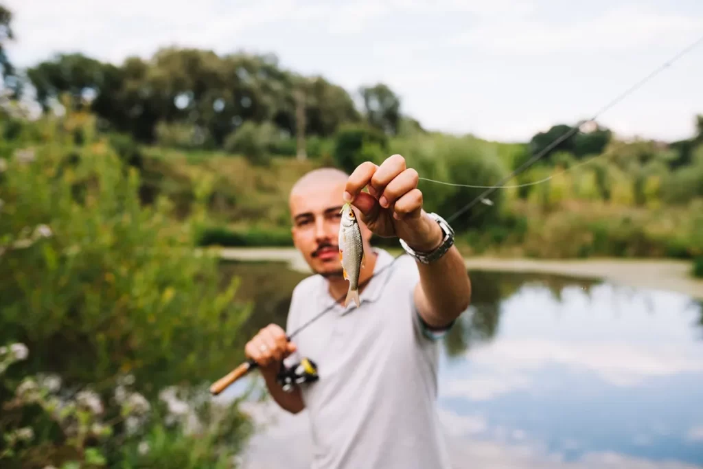 Homem segurando um pequeno peixe recém-pescado em um ambiente natural com lago ao fundo.