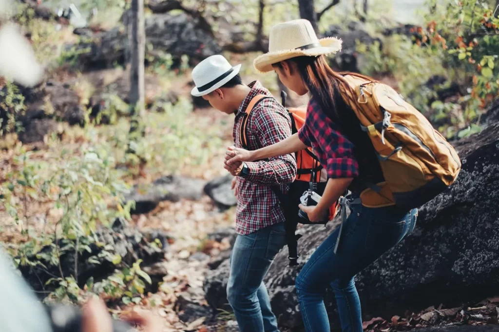 Casal com roupas de trilha e mochilas caminhando em meio à natureza, explorando uma trilha com vegetação e rochas, representando o turismo rural e ecológico.