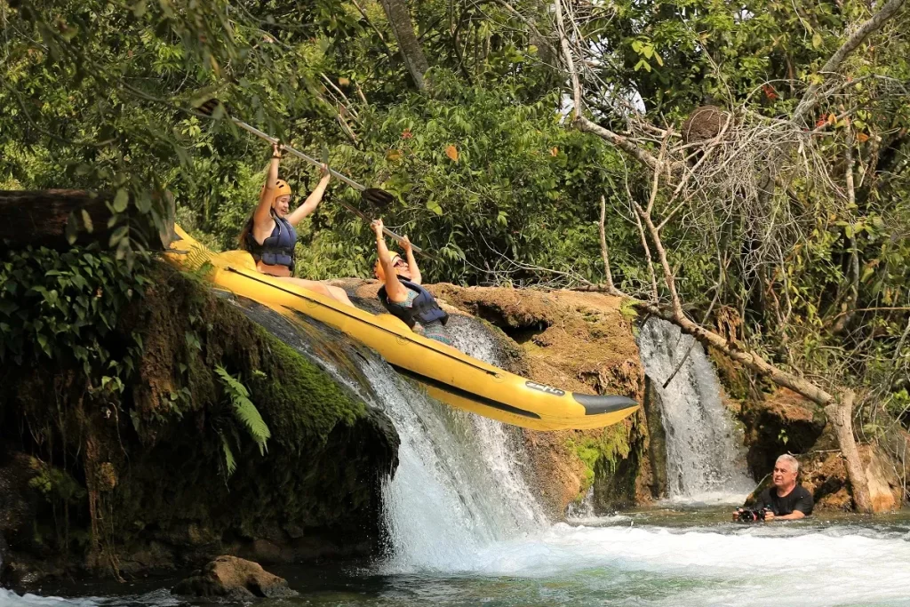 Duas pessoas em caiaque amarelo descendo um pequeno salto de cachoeira cercado por vegetação, enquanto um fotógrafo registra a cena na água.
