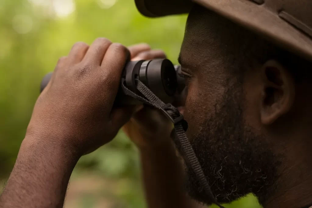 Homem com chapéu de explorador usando binóculos para observar a natureza em meio a um ambiente verde.