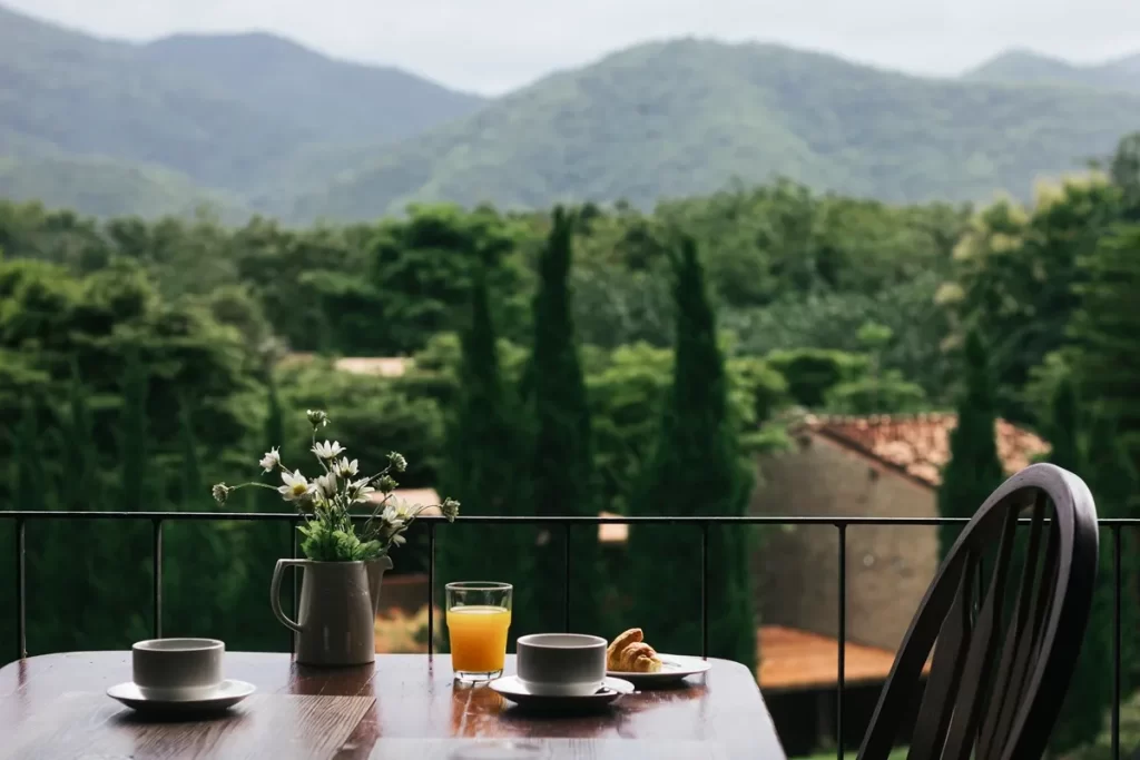 Mesa rústica em varanda com café da manhã servido, incluindo xícaras, suco de laranja e croissant, com vista para montanhas e vegetação exuberante.