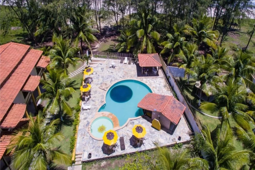 Área de piscina cercada por guarda‑sóis amarelos e palmeiras, com chalés de telha vermelha e praia ao fundo