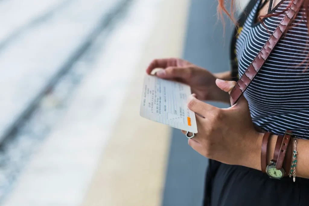 Mulher segura uma passagem de trem enquanto espera na plataforma de uma estação ferroviária, com trilhos visíveis ao fundo.