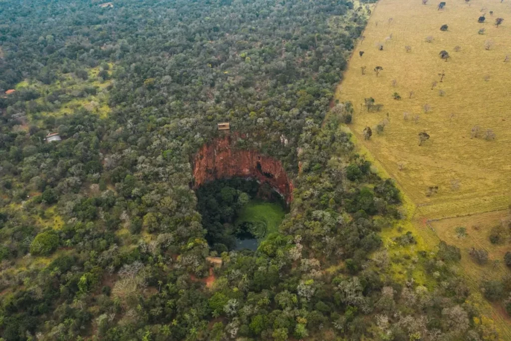 Vista aérea de uma dolina em Bonito (MS), com paredes de pedra avermelhada e fundo verde de vegetação ao redor de uma lagoa.