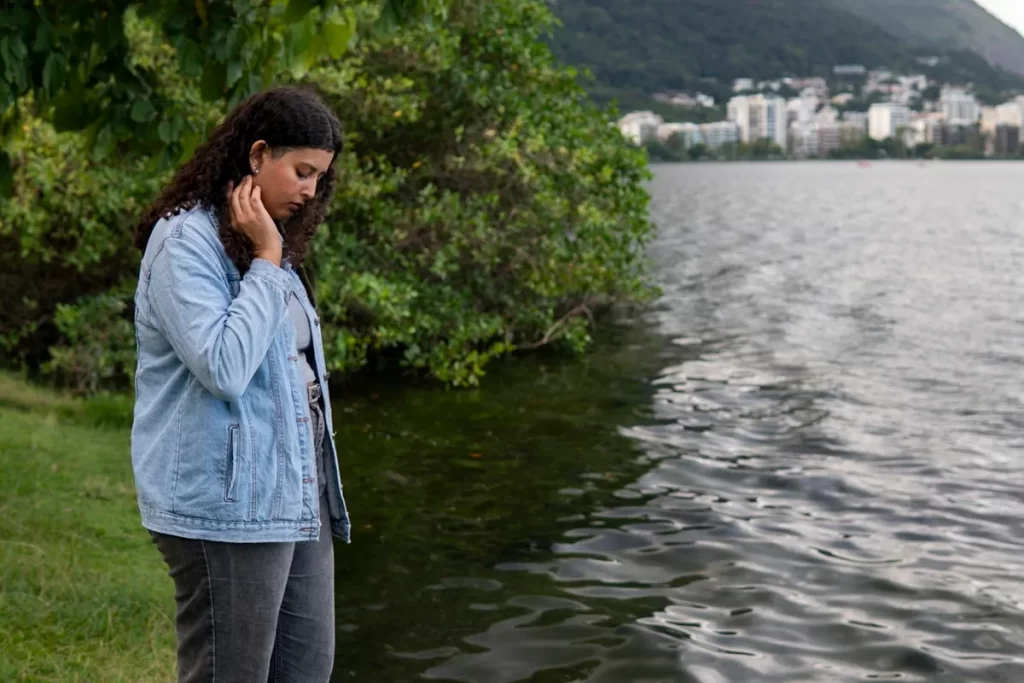 Mulher com expressão pensativa olhando para a água de um lago, em pé na margem cercada por vegetação, com cidade e morro ao fundo