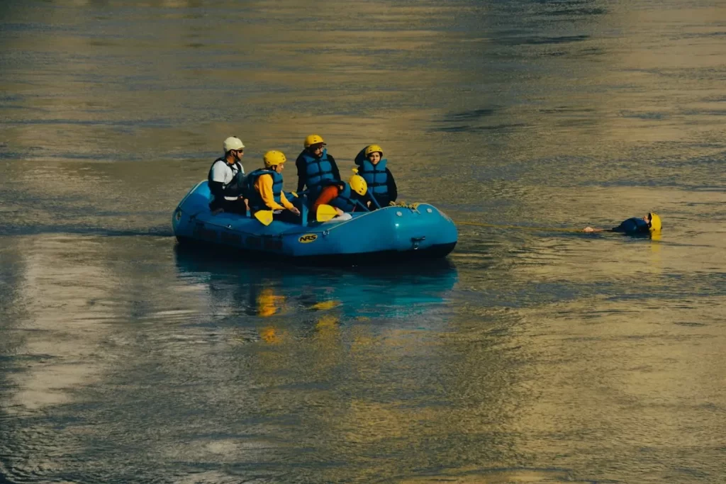 Grupo de pessoas em bote inflável azul puxando com corda uma pessoa com capacete flutuando no rio
