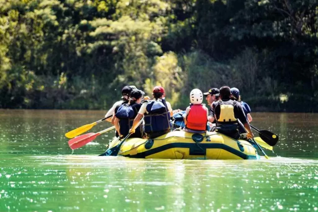 Turistas com coletes salva-vidas remando juntos em bote inflável amarelo sobre rio calmo cercado por vegetação