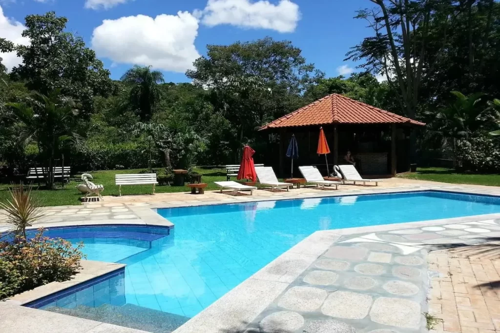 Piscina ao ar livre com espreguiçadeiras, guarda-sóis e quiosque coberto em área cercada por vegetação nativa
