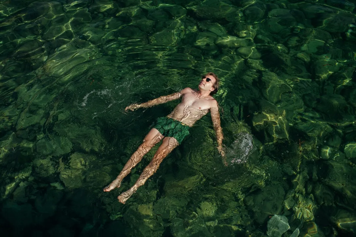 Homem flutuando de costas em lagoa de água cristalina, relaxando sob o sol.