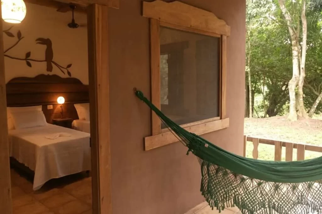Vista de um quarto de pousada com camas feitas, arte de tucano na parede e varanda com rede verde e vista para a natureza.