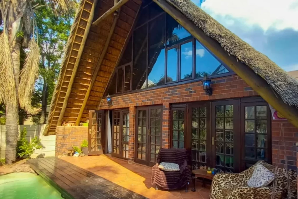 Fachada de uma casa de campo com telhado de palha íngreme, paredes de tijolo, grandes janelas de vidro e um deck de madeira próximo a uma piscina.