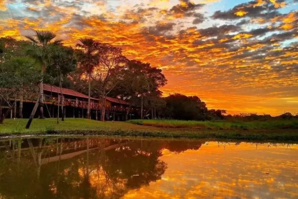 Vista panorâmica de uma lodge à beira de uma lagoa, com um pôr do sol alaranjado e avermelhado espetacular refletindo na água e no céu com nuvens dramáticas.