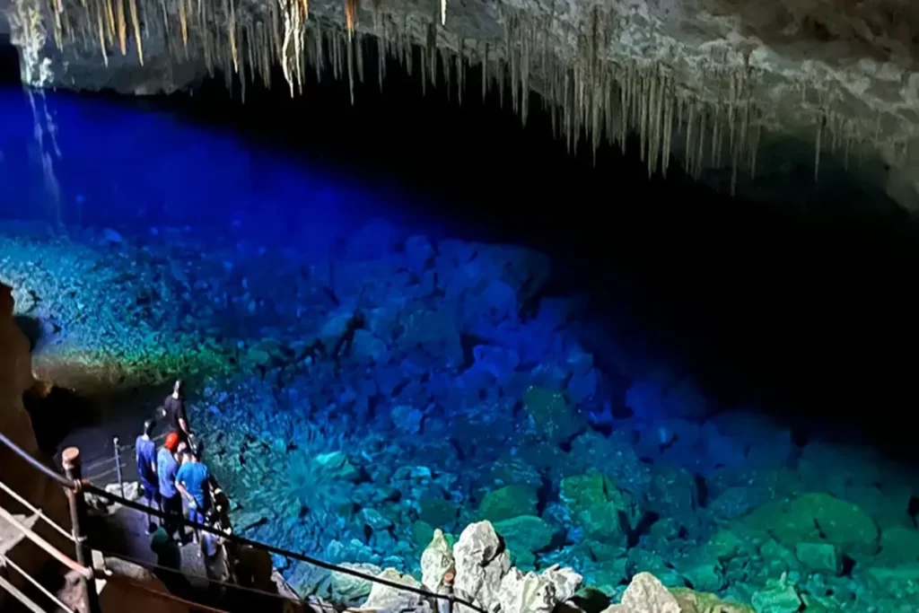 Visão aérea da Gruta do Lago Azul em Bonito, Mato Grosso do Sul, Brasil, com um deslumbrante lago de águas azuis profundas em uma caverna escura, estalactites penduradas no teto e turistas observando da beira.
