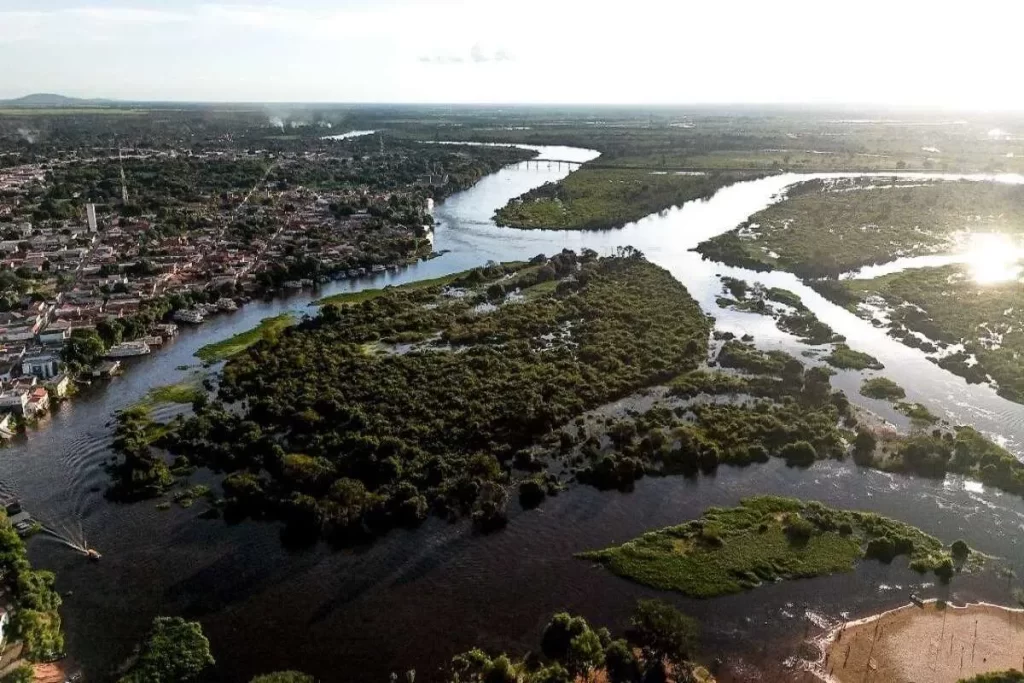 Vista aérea panorâmica de um vasto rio com suas águas escuras e afluentes que serpenteiam por uma paisagem exuberante, contornando ilhas verdes densamente arborizadas. À esquerda, uma cidade com arquitetura antiga margeia o rio, enquanto à direita, a vegetação densa se estende até o horizonte, com o reflexo do sol na água em alguns pontos. Uma pequena lancha é visível na parte inferior esquerda do rio.