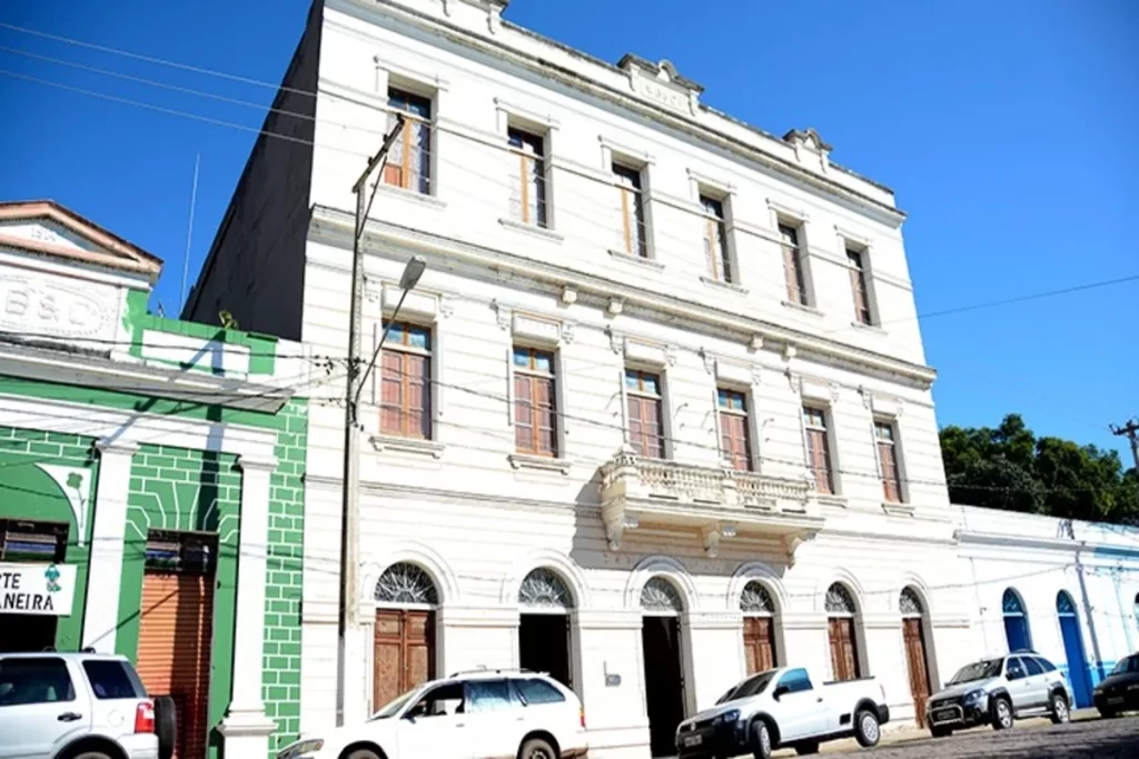 Vista de uma rua em Corumbá, Mato Grosso do Sul, com um grande edifício branco de arquitetura histórica em destaque e outros prédios coloridos ao lado, sob um céu azul. A imagem representa o patrimônio cultural e arquitetônico da cidade.