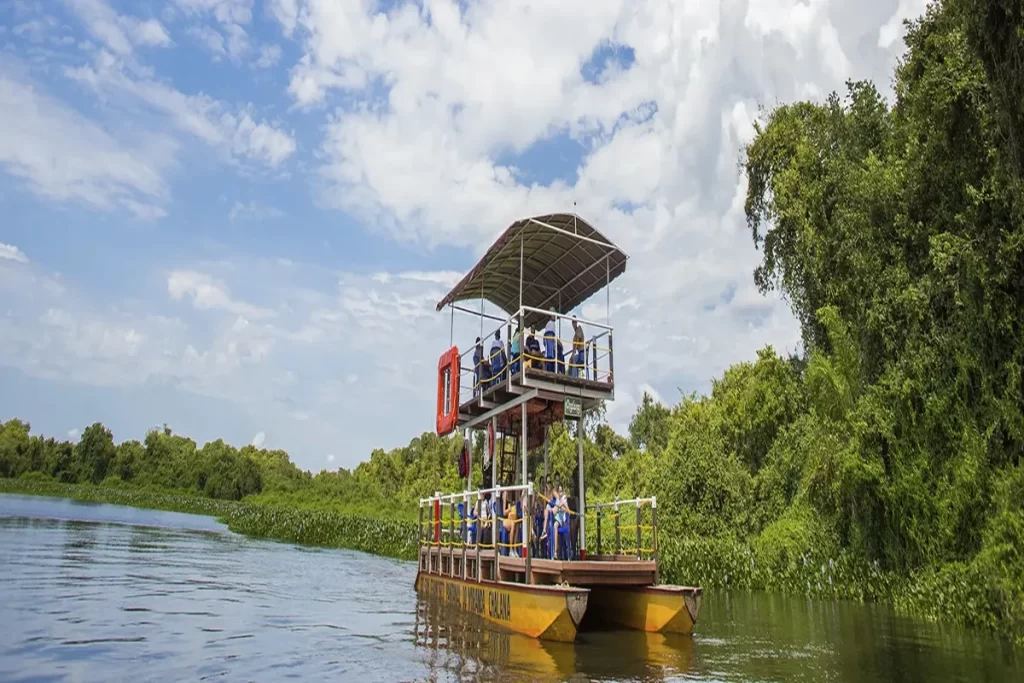 Chalana navegando em rio no Pantanal com turistas, apreciando a paisagem natural exuberante sob céu azul com nuvens.