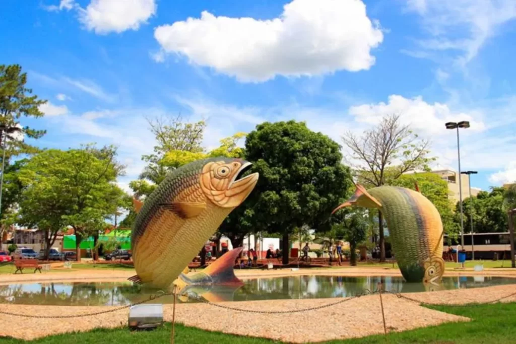 Esculturas de peixes em uma praça na cidade de Bonito, Mato Grosso do Sul, sob um céu azul com nuvens brancas.