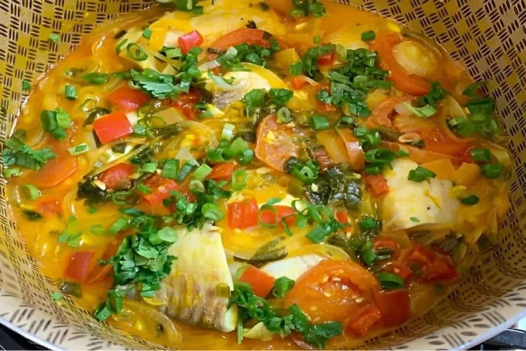 Uma panela com moqueca de peixe decorada com rodelas de tomate e cebola e coentro fresco picado. A imagem evoca o sabor rico e o frescor do prato, com a textura do recipiente de bambu entrelaçado.