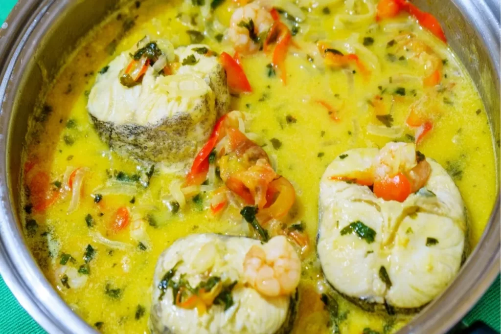 Uma panela de moqueca com peixe branco, camarões e vegetais. A imagem mostra a consistência e as cores vibrantes do prato, com seus ingredientes principais: peixe, camarão, pimentão e coentro.