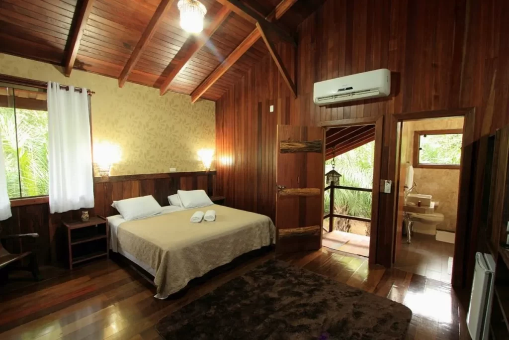 Interior de cabana de madeira com cama queen size, teto abobadado, ar condicionado e acesso à varanda, suíte de hotel cabanas confortável.