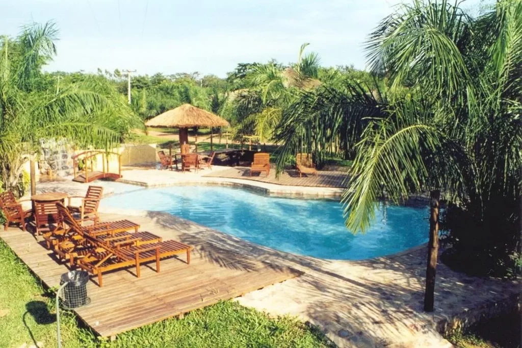 Piscina em formato orgânico com deck de madeira e palmeiras, bangalô de palha com mesas e cadeiras de madeira, ideal para lazer em hotel cabanas tropical.