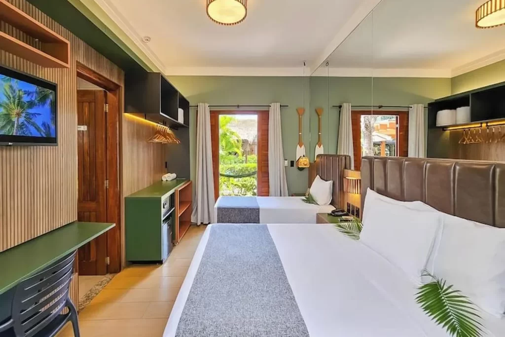Interior de um quarto de hotel ou pousada com duas camas, decoração moderna em tons de verde-oliva, painéis de madeira ripada e janelas com vista para o jardim, representando a acomodação de luxo da Pousada Arte da Natureza em Bonito.