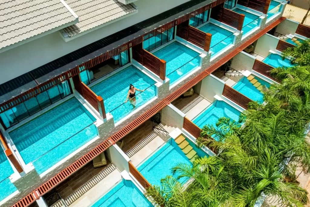 /Visão de cima (aérea) de um hotel ou pousada de luxo com arquitetura moderna e telhado branco, apresentando várias suítes enfileiradas, cada uma com sua própria piscina individual de azulejos azuis e cercada por madeira. Uma mulher de maiô escuro é vista na piscina central. Palmeiras tropicais em primeiro plano destacam o ambiente em Bonito, Mato Grosso do Sul.