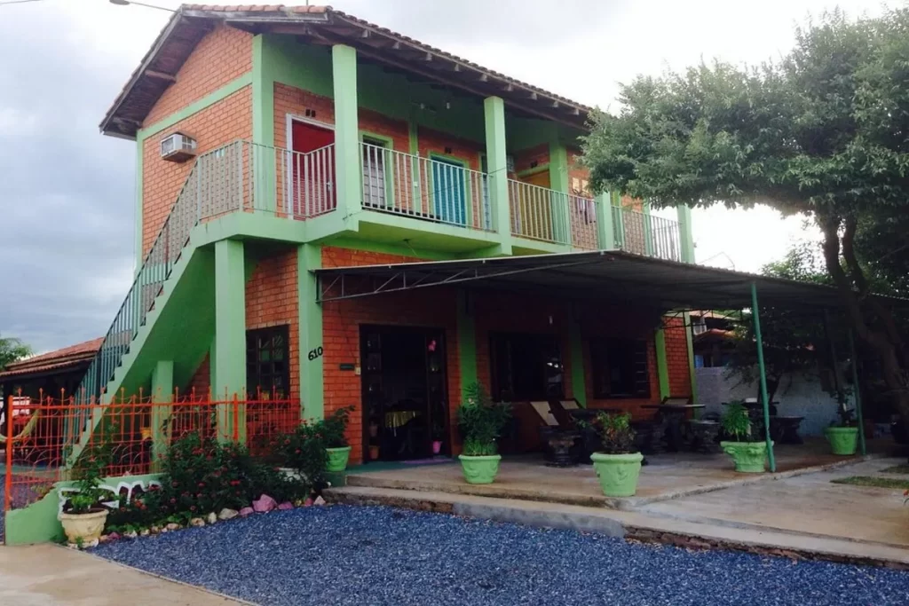 Fachada da Pousada Primavera, prédio de dois andares com tijolos aparentes, varanda e jardim com vasos de plantas.