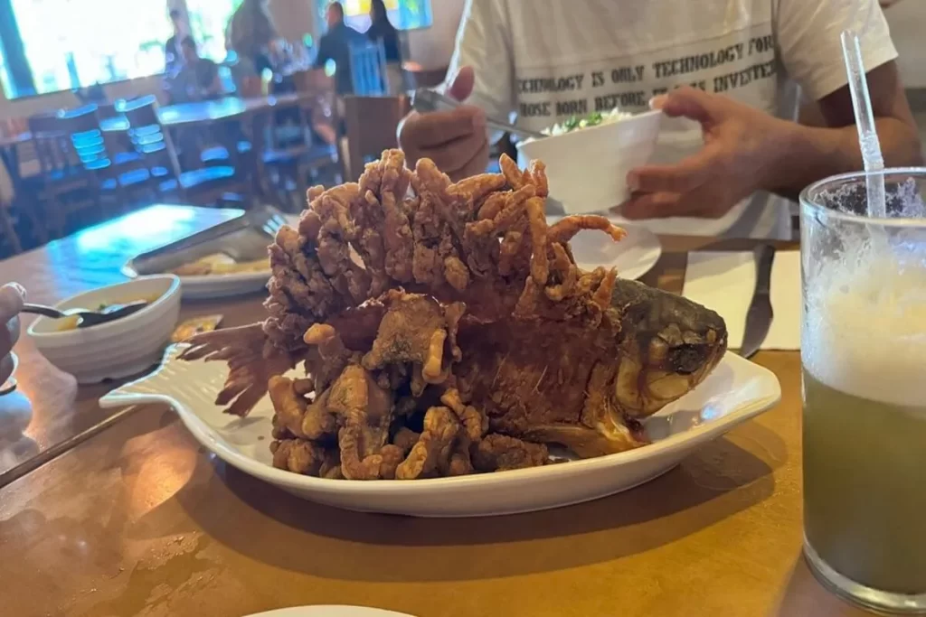 Um prato em forma de peixe com um peixe inteiro frito, crocante e dourado, acompanhado por um copo de suco e uma tigela de arroz. A imagem destaca a textura crocante e o peixe em um prato branco sobre uma mesa de madeira.