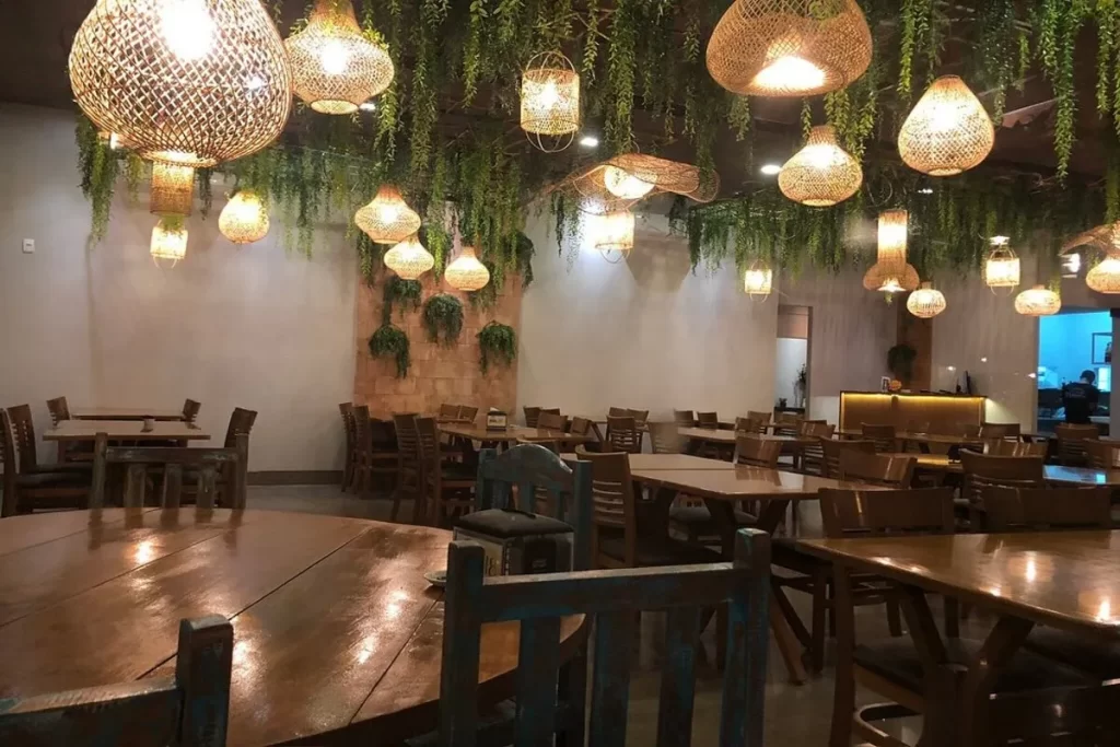 Interior do Varandas Restaurante com mesas e cadeiras de madeira, iluminação quente de luminárias artesanais penduradas no teto, e folhagem verde luxuriante adornando o ambiente.