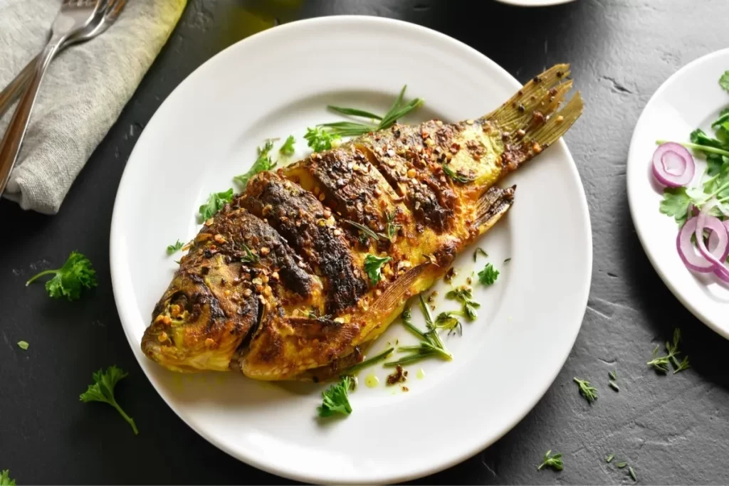 Um pacu assado inteiro, com a pele dourada, servido em um prato branco sobre uma superfície escura, guarnecido com ervas frescas e pimenta calabresa.