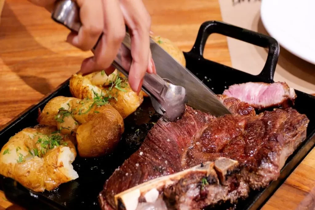 Um corte grosso de carne vermelha suculenta sendo fatiado em uma chapa de ferro, acompanhado por batatas assadas, prato do restaurante Bacuri Cozinha Regional.