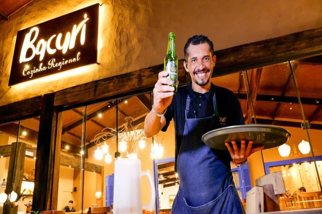 Garçom sorridente segurando uma garrafa de cerveja e uma bandeja, em frente à fachada iluminada do restaurante Bacuri Cozinha Regional em Bonito, MS.