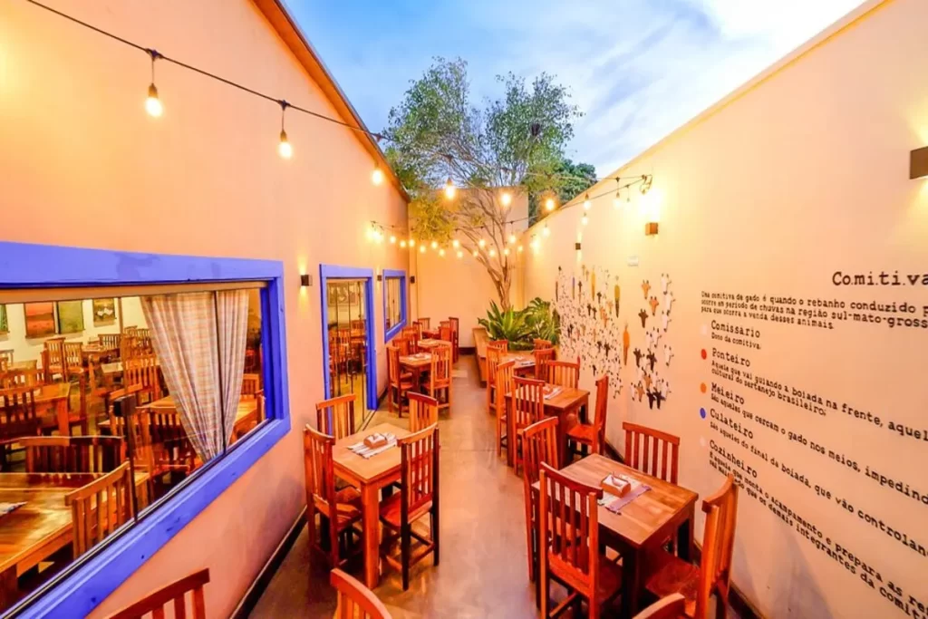 Pátio externo de restaurante com mesas e cadeiras de madeira, paredes em tom pêssego, iluminação com luzes de gambiarra e painel temático sobre a Comitiva da pecuária sul-mato-grossense.