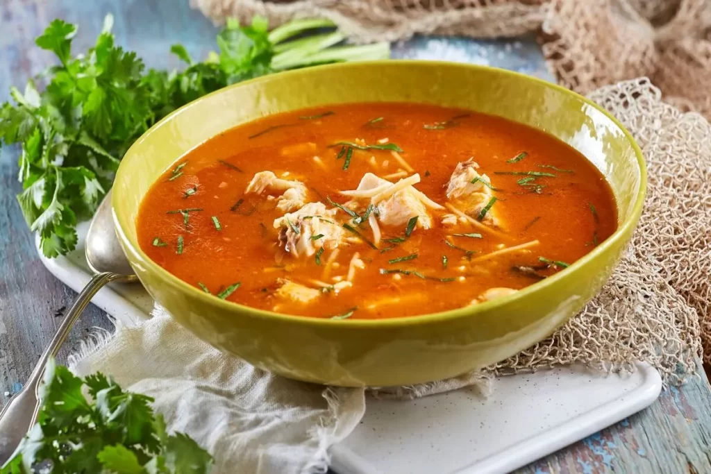 Uma tigela verde cheia de sopa de peixe de cor alaranjada, com grandes pedaços de peixe e guarnecida com ervas picadas.
