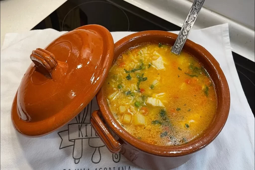Uma sopa de peixe quente e colorida, com pedaços de peixe, batata e ervas, servida em uma panela de barro tradicional, com a tampa ao lado.