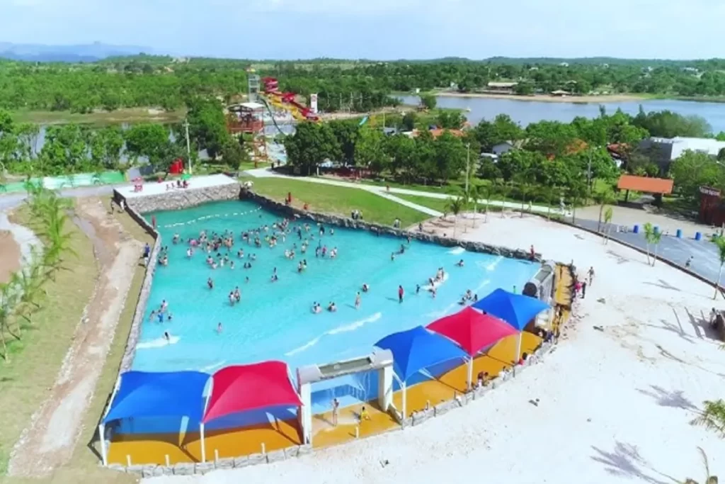Vista aérea de uma grande piscina de ondas em um parque aquático, cheia de pessoas se refrescando. O Acqua Park Pantanal apresenta toldos de sombra coloridos (azul, vermelho e amarelo) na borda, com vegetação e um lago natural visível ao fundo, destacando o contraste entre a diversão artificial e a natureza.