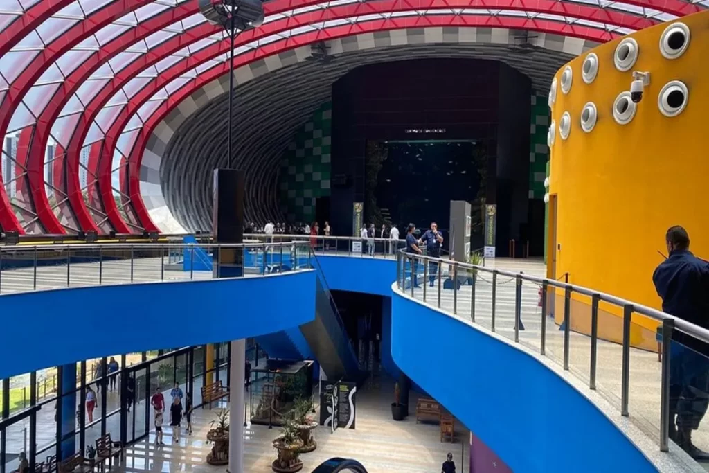 Interior do Bioparque Pantanal, em Campo Grande, com arquitetura curva e futurista, pilares vermelhos e corrimãos azuis, mostrando a área de circulação e tanques de aquário ao fundo.