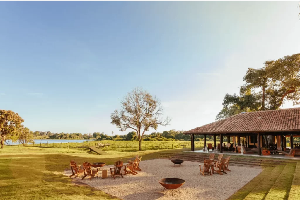 Vista da área de convívio do Refúgio Ecológico Caiman no Pantanal, com cadeiras de madeira rústicas em um pátio, gramado e lagoa ao fundo sob o céu azul.