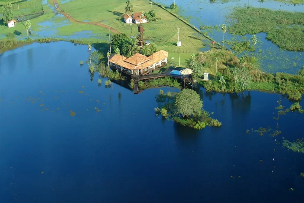 Vista aérea de uma fazenda pantaneira (lodge) com telhado de terracota, cercada por grandes áreas de água e campos alagados sob um céu azul. A imagem ilustra o clima do Pantanal durante a estação de cheia, com a vegetação verdejante contrastando com o azul intenso do lago.