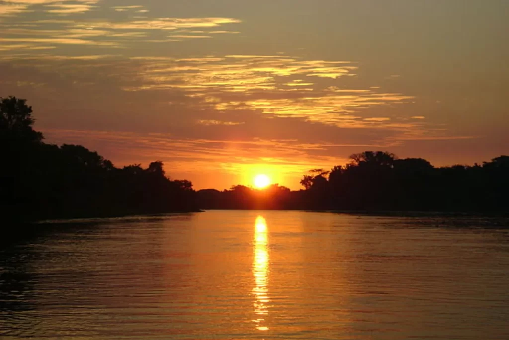 Cenário de um pôr do sol deslumbrante sobre um rio calmo no Pantanal, com o céu em tons de laranja e dourado. O sol está baixo na linha do horizonte, refletindo intensamente na água, e a margem é marcada pela silhueta da vegetação pantaneira.
